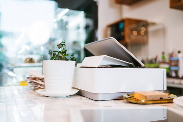 A modern point-of-sale terminal sits on a countertop next to a coffee cup and a small potted plant in a bright, stylish cafe setting.