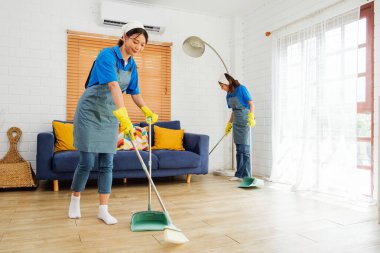 Two uniformed cleaning professionals sweep and maintain a modern living space with wooden floors and a blue sofa, showcasing efficient, reliable facility care in a professional service setting.