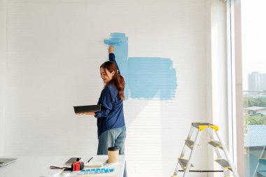 Gülümseyen bir kadın, neşe ve yaratıcılığını ifade eden parlak ve modern bir odada duvarı maviye boyar. Bu görüntü kişisel gelişimi, iç dönüşümü ve ev dekorasyonunun ruhunu yakalar..