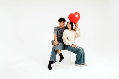 Neşeli bir çift, temiz bir stüdyoda birbirine yakın oturuyor, gülümsüyor ve ellerinde kırmızı kalp şeklinde bir balon tutuyorlar. Aşkı, mutluluğu ve sevgiyi simgeliyor. Sevgililer Günü ve yaşam tarzı görselleri için mükemmel.