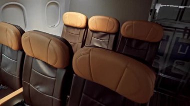 Ticari bir jet kabininin penceresinin yanındaki kahverengi ve siyah deri uçak koltukları, boş ve biniş için hazır, başlıkları ve kollukları görünür..