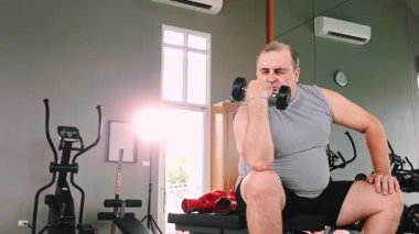 Kilolu bir insan spor salonundaki bir bankta halterle biseps kıvırıyor, kardiyo makineleri ve ağırlık çalışmalarına odaklanıyor..