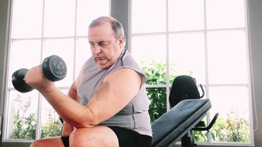 Spor salonunda oturan yaşlı, kilolu bir adam bir bankta halterli bir kıvrım yapıyor. Egzersiz ekipmanları ve parlak pencere ışığı arasında güç eğitimine odaklanıyor..