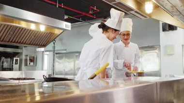 Ticari bir mutfakta iki aşçı, biri diğerine öğretirken küçük bir tencereyi paslanmaz çelik başlıklar ve tezgahların altında karıştırıyor.