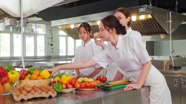 Profesyonel bir mutfakta üç aşçılık öğrencisi, bir eğitmenin rehberliğinde renkli taze meyve ve yumurtaların yanında soğan hazırlığı yapmayı öğreniyor..