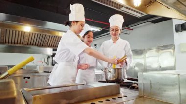 Profesyonel bir mutfakta aşçılar gülümserken, bir eğitmen öğrencilere fırında demlik karıştırma konusunda rehberlik ediyor..