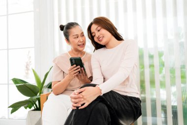 İki kadın. Anne ve kız akıllı telefondan bir şey gösterilirken evde otururlar; rahat bir sohbet ve pencere perdeleri ve saksısı olan parlak bir odada gülümserler..