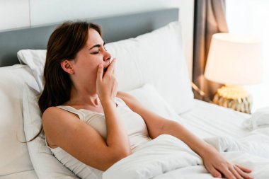 Uykulu kadın beyaz bir battaniyenin altında yatarken esniyor. Yatağın başucundaki lambadan sabah ışığı parlıyor..