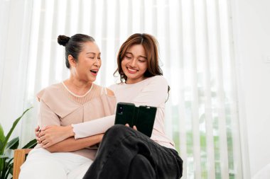 İki kadın. Anne ve kız evde oturuyor, gülümsüyor ve sarılıyor ve parlak bir pencerenin yanında dikey perdelerle akıllı bir telefona bakıyor..