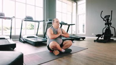 Modern bir spor salonunda, ayak değirmenleri ve kardiyo makineleriyle çevrili bir yoga minderinde meditasyon yapan bir adam, sakin, güneşli bir farkındalık ve sağlığın tadını çıkarıyor..