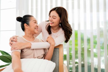 Anne ve kızı. Genç bir kadın evde yaşlı bir kadına sarılıyor, parlak doğal ışıkta sıcak ve neşeli bir anı paylaşıyor..