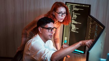 İki yazılım geliştiricisi ikili monitörlerde işbirliği yaparak, loş bir ofiste kodu gözden geçirir; biri ekranı işaret ederken diğeri dikkatle gözlemler.