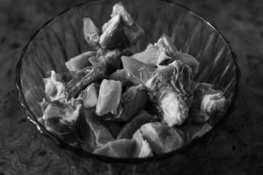 A black and white, slightly elevated close-up of raw, diced meat in a glass bowl. The monochrome tone emphasizes the textures and shapes of the uncooked protein.