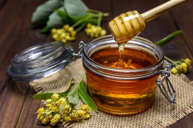 Linden balı, ıhlamur çiçeklerinin yanındaki kavanoza tahta sopayla dökülüyor. Seçici odak.