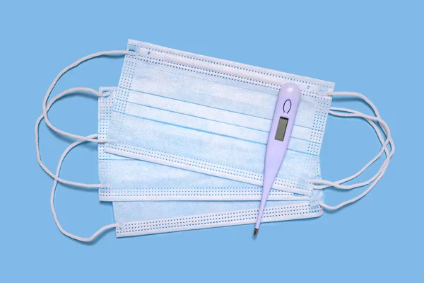 Tıbbi koruyucu yüz maskeleri ve insan vücudunun açık mavi arka plandaki sıcaklığını ölçmek için dijital termometre..