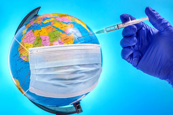 A doctor\'s hand in a glove injects a syringe with the coronavirus vaccine into a earth globe covered with a surgical face mask. Concept of the world\'s fight against COVID-19. Selective focus.