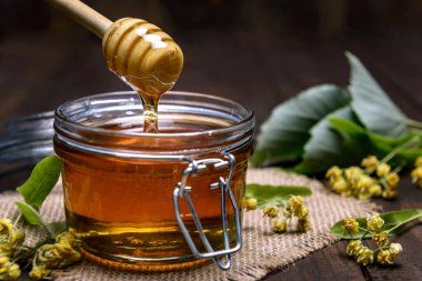 Linden balı, ıhlamur çiçeklerinin yanındaki kavanoza tahta sopayla dökülüyor. Seçici odak.