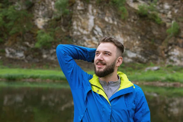Portrait of happy smiling bearded man in jacket with hand thrown back behind head looking up at natural background of rocky shore. Satisfied with life. Dreamy look. Tourist admires beautiful landscape