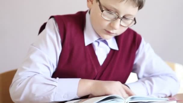 Niño con gafas leyendo un libro mientras está sentado en un escritorio — Vídeo de stock