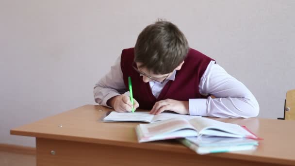 Хлопчик в окулярах пише в блокноті, сидячи за столом — стокове відео