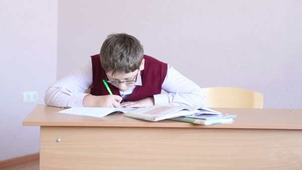 Boy with glasses writes in a notebook sitting at a desk — Stock Video