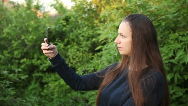 Młoda dziewczyna robi selfie stojący w parku, w pobliżu drzewa — Wideo stockowe