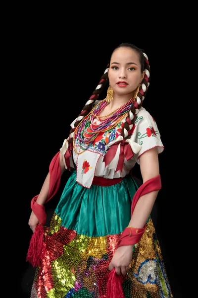 Latin woman dressed as a china poblana with a skirt embroidered in colored sequins, a red shawl, braided bows and a smiling, black background