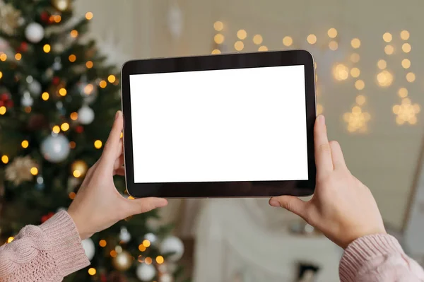 Mock up, tablet in the hands of a girl. Against the background of New Year and Christmas holidays, a cozy interior with a decorated Christmas tree