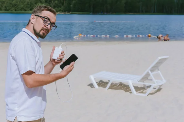Gözlüklü bir adamın portresi sahildeki bir elektrik bankasından akıllı telefonu şarj ediyor. Arkaplanda kum, güneş ve su var.