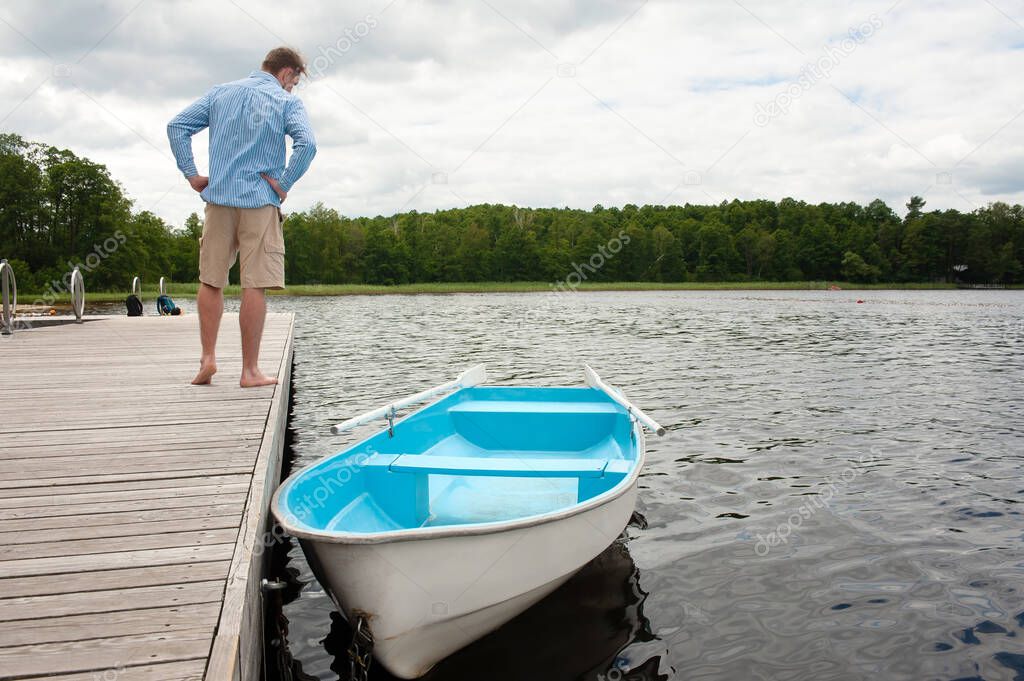 A man standing on a wooden pier looks at a boat with oars in the water