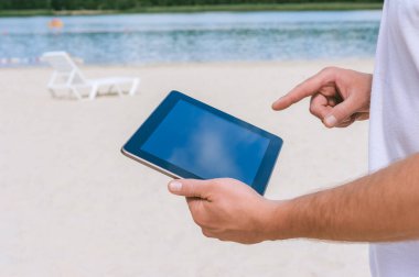 Sahilde bir adam parmağını tablet ekranın önünde tutuyor. Kum ve suyun arka planına karşı