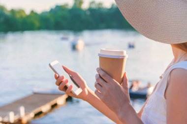 Yakın plan, elinde bir kupa kahveyle bir kızın elinde akıllı telefon. Arka planda su, yat, tekne ve katamaranlarla dolu bir set var.