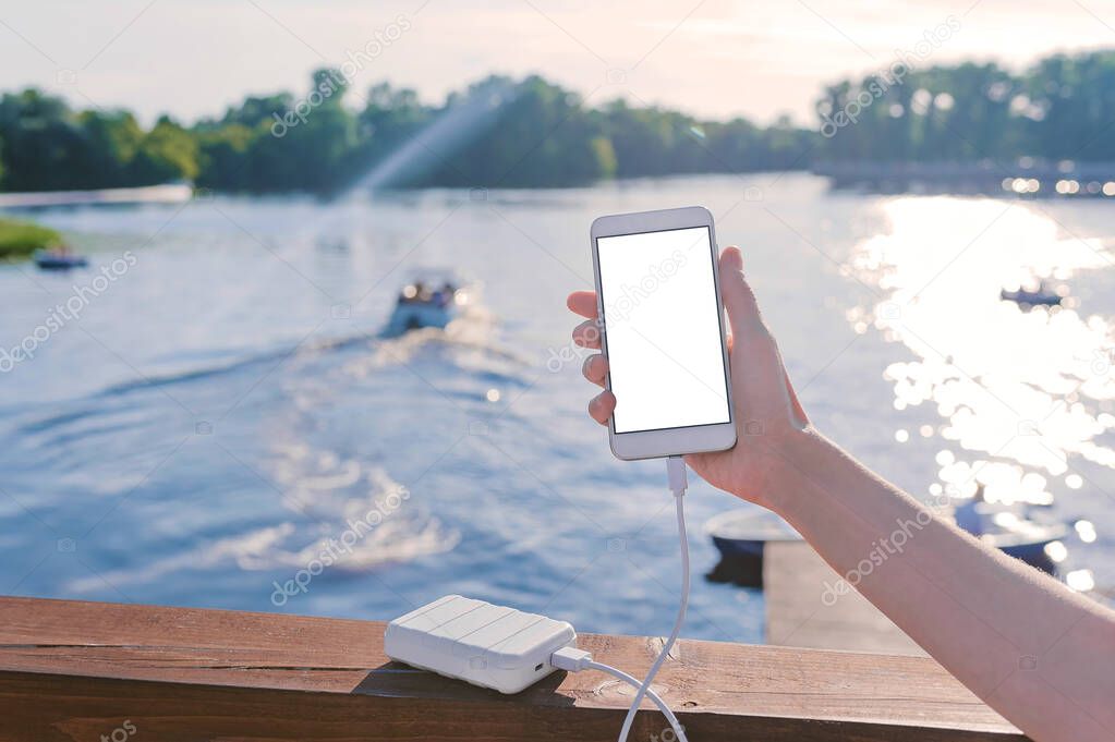 Mock up of a smartphone in the hand of a girl on the pier. Charge your phone with Power Bank. Against the background of the river, lake with a boat