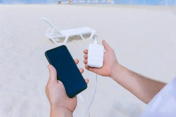 Power Bank Energy charges the phone into the hands of a man on the beach. Against the backdrop of sand and a sun lounger