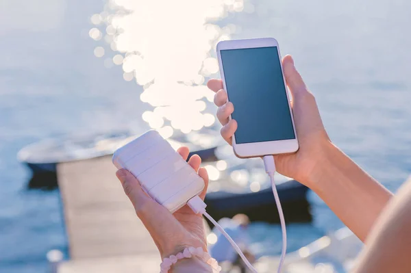 Rıhtımdaki bir kızın elinde akıllı telefon. Telefonunu Power Bank ile şarj et. Bir nehrin, gölün, köprünün ve teknenin arka planında.