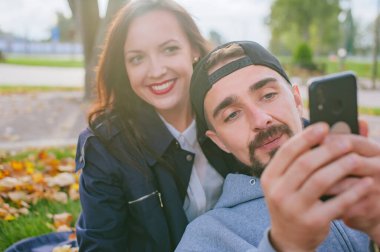 Elinde akıllı telefon olan bir adam sonbaharda yaprakların arasında sevgili mutlu kızıyla oturuyor.