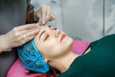 Kozmetik uzmanı, kanepede yatan genç bir kızın yüzüne iğne yapıyor.