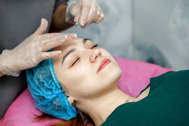 Kozmetik uzmanı, kanepede yatan genç bir kızın yüzüne iğne yapıyor.