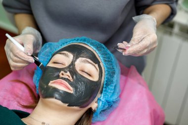 Güzellik uzmanı, fırçayla genç bir kızın yüzünü soymak için siyah bir maske takıyor. Bir kozmetik uzmanı, genç bir kızın kanepede yatması için deri yüzme prosedürü uygular..