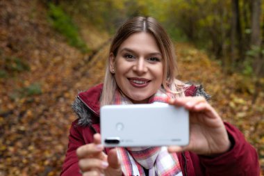 Güzel genç bir kadın akıllı telefonuyla çevrimiçi içerik oluşturuyor. Renkli bir sonbahar geçmişi var.