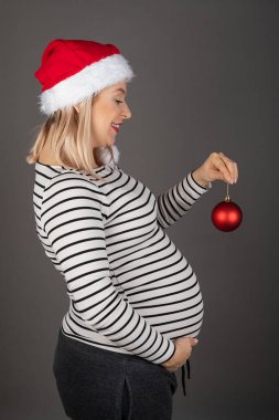 Üçüncü ayda hamile bir kadın gri arka planda Noel aksesuarları takıyor.