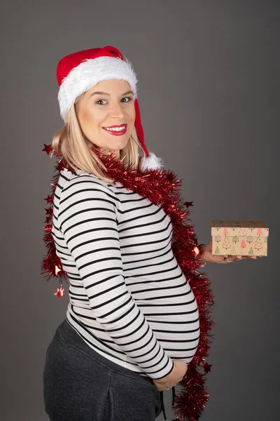 三个月前 漂亮的孕妇头戴圣诞帽 头戴灰色背景的红色圣诞花环 手里拿着一件礼物 — 图库照片
