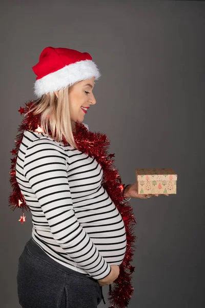 三个月前 漂亮的孕妇头戴圣诞帽 头戴灰色背景的红色圣诞花环 手里拿着一件礼物 — 图库照片
