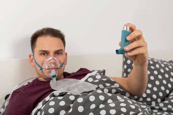 Male patient with oxygen mask at home is lying in bed, holding an inhaler spray with salbutamol