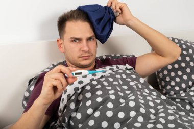 Hasta, beyaz bir adam elinde dijital termometreyle evde yatıyor.