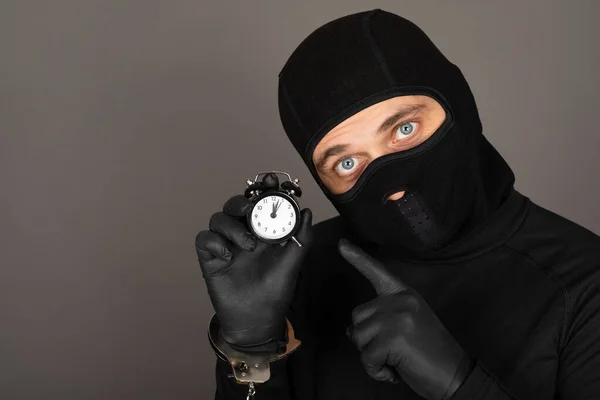 黒いマスクと強盗の容疑者の衣装を着た若い男の写真 灰色の背景の前に手錠を身に着けています — ストック写真