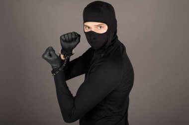 Siyah takım elbiseli ve yüzünde maske olan hırsız elleri kelepçeli poz veriyor.