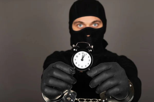 黒いマスクと強盗の容疑者の衣装を着た若い男の写真 灰色の背景の前に手錠を身に着けています — ストック写真