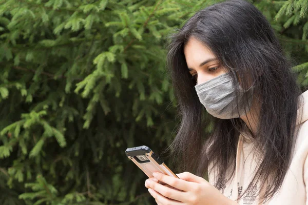 The girl in the medical mask looks at the phone. A woman with long black hair holds a mobile phone. Health care, covid19. The wearing of masks in public places. The girl smiles in a medical mask.