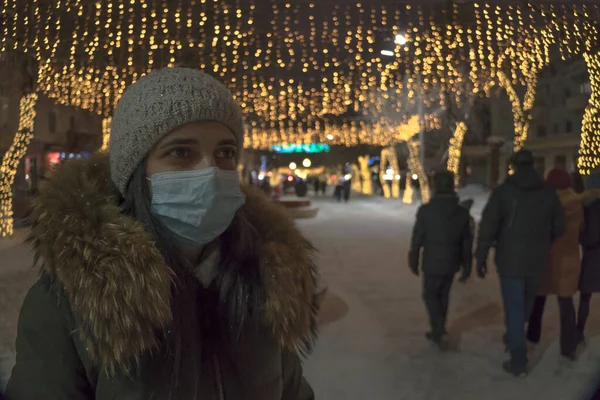 Yılbaşı sokağında maskeli bir kız. Yüzü maskeli bir kadın Noel ve yeni yıl için süslenmiş dükkanları olan bir caddede duruyor. Grip salgını sırasında tatiller. Güvenli Xmas ve yeni yıl
